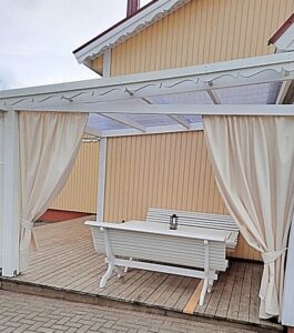 Šviesi kreminė terasa, pieno spalvos terasos užuolaidos, dažyti mediniai lauko baldai, baltas lauko suolas.